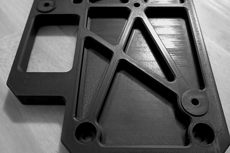 Black additively manufactured mounting plate with diagonal stiffening ribs and pockets.