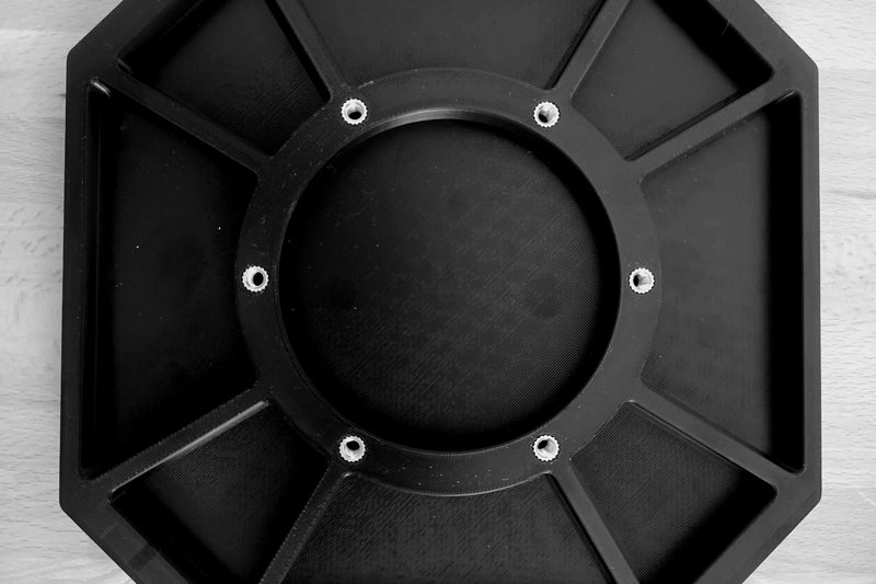 Black octagonal additively manufactured plate with radial ribs, central bore and brass threaded inserts.